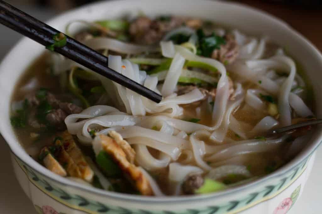 Vietnamese beef noodle soup Pho Bo with garnish of leaves of cilantro, Asian basil and bean sprouts on kitchen table. Young woman is cooking in background. Traditional healthy food.