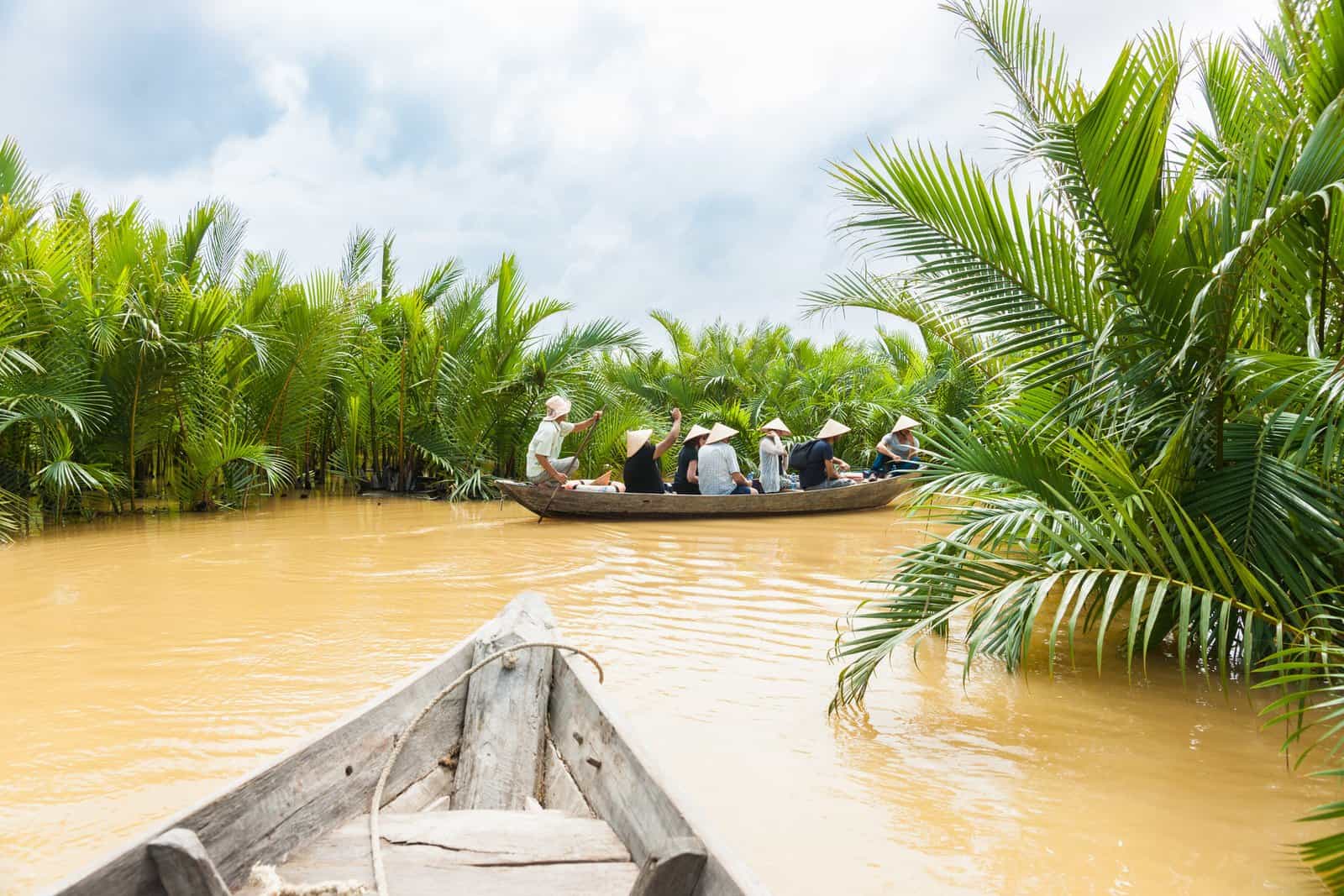 River scene with Group of tourists are poled along river between luxuriant palm lined sides in old traditional wooden canoe