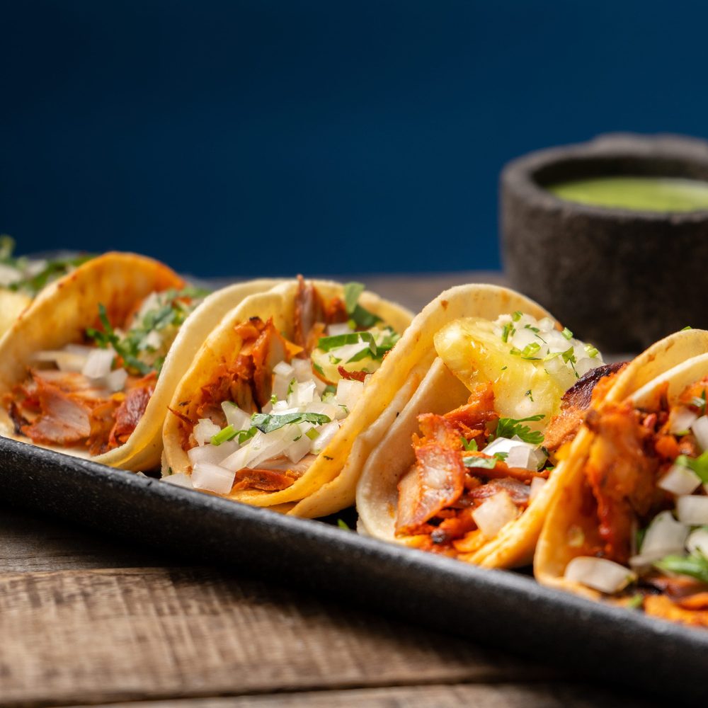 Tacos al pastor with pineapple, onion and coriander on a metal tray beside a bowl of salsa verde