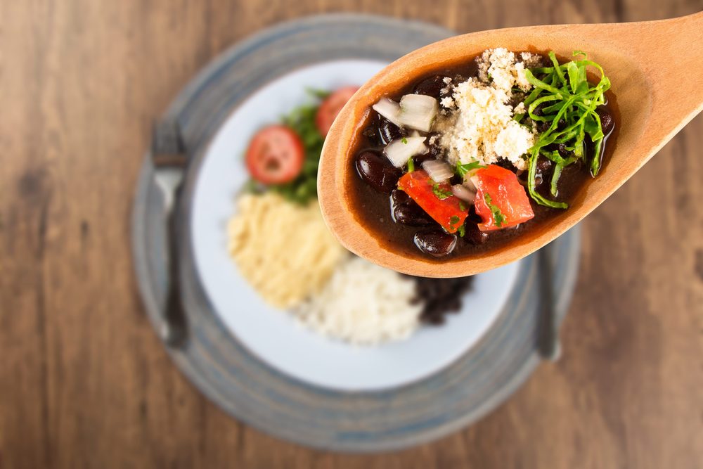 A wooden spoon of feijoada, the black bean stew, over a plate with rice, farofa and collards